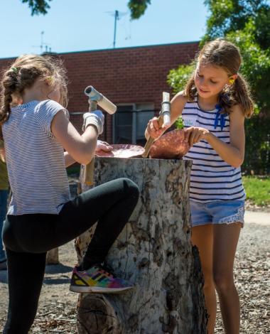 Two children standing next to each other hammering copper bowls, their work surface is a tree stump, they are working outdoors in the sunshine. There is a red brick building, a leafy green tree and the blue sky in the background.