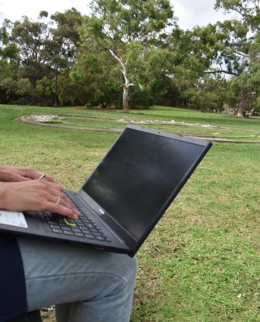 A person sits with a laptop on their lap and Gasworks Park in the background - grass, trees and blue sky