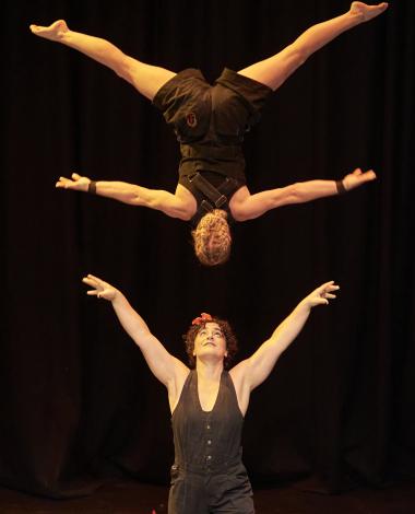 A performer looks up, arms raised and open wide. Another performer is upside down in mid air. They are looking at each other, one is smiling.