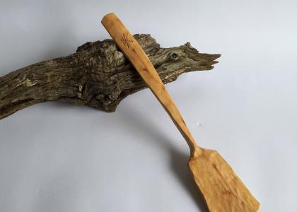 A hand carved spatula resting on a piece of wood. 