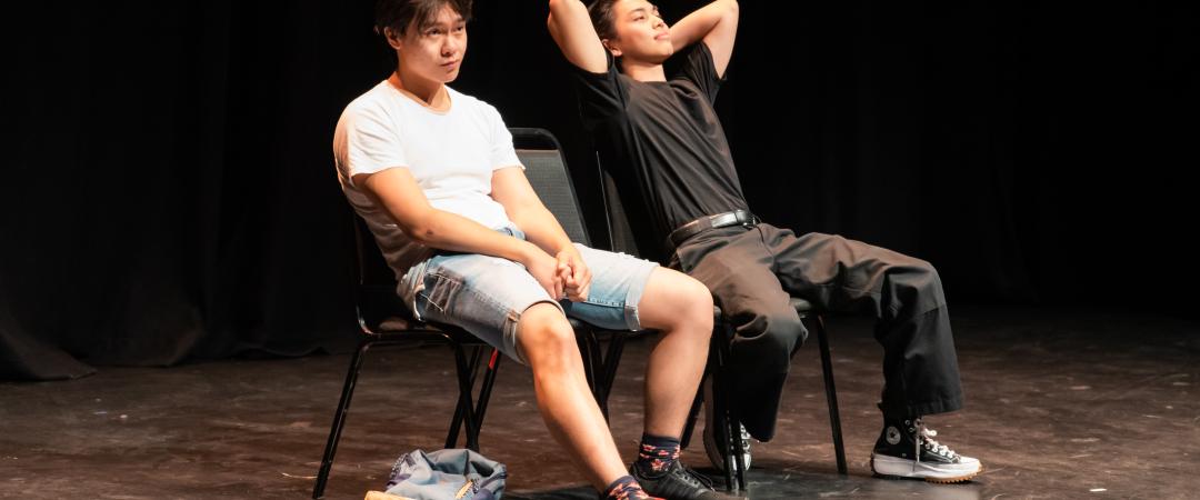 Two casually dressed young Asian men sit next to each on black chairs in a theatre with a black stage floor and a black curtain behind them.