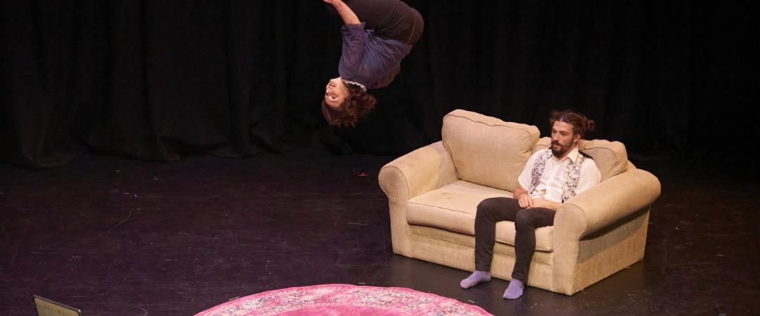 A performer is sitting on a couch - he looks straight ahead, but does not seem to notice the performer doing a backflip in mid air right in front of him; he is too focused watching the laptop that is in his direct eyeline. There is a round Persian rug ion the ground between them.
